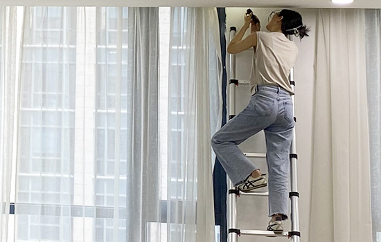 Une fille grimpe sur une échelle télescopique en131 pour réparer des rideaux